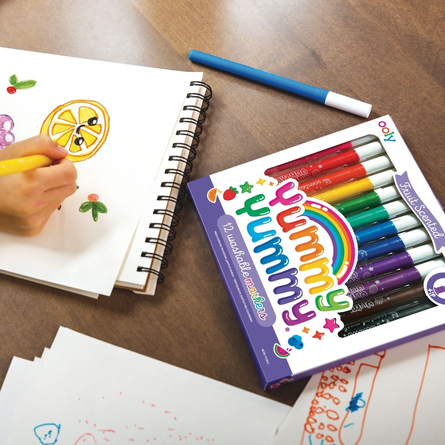 A set of 12 Yummy Yummy scented markers by ooly, with pointed tips and a variety of colors, including black, red, orange, yellow, green, blue, and purple, packaged in a clear plastic case with a colorful label.