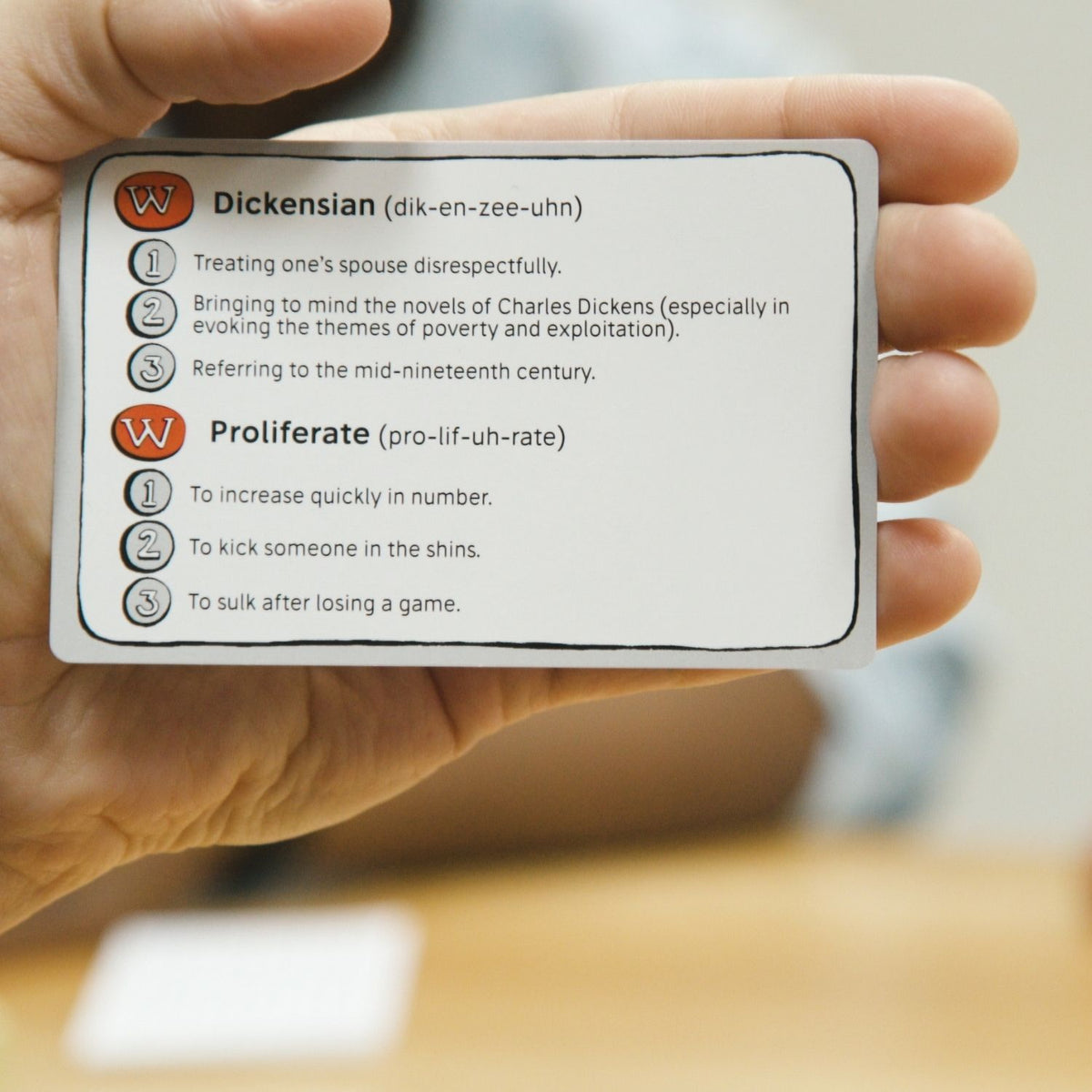 Hand holding a card with definitions of words like 'Dickensian' and 'Proliferate'.