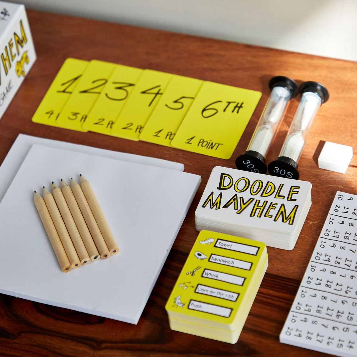 Doodle Mayhem game materials including pencils, a whiteboard, and a timer on a wooden surface.