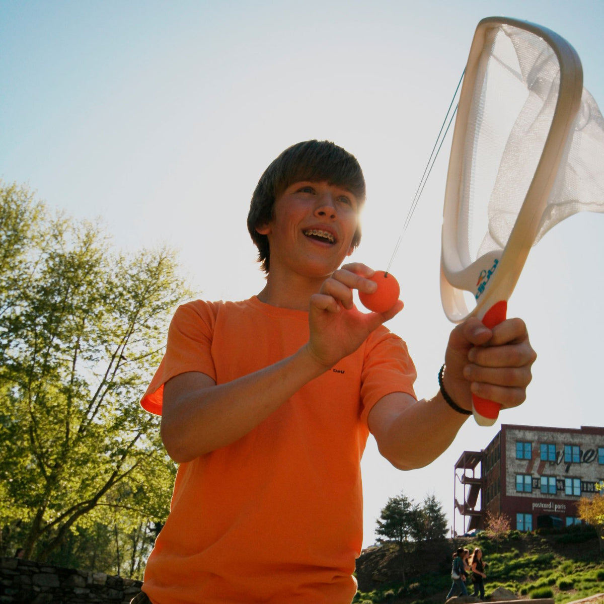 Person in orange shirt holding a SlingBall net outdoors with trees and building in the background