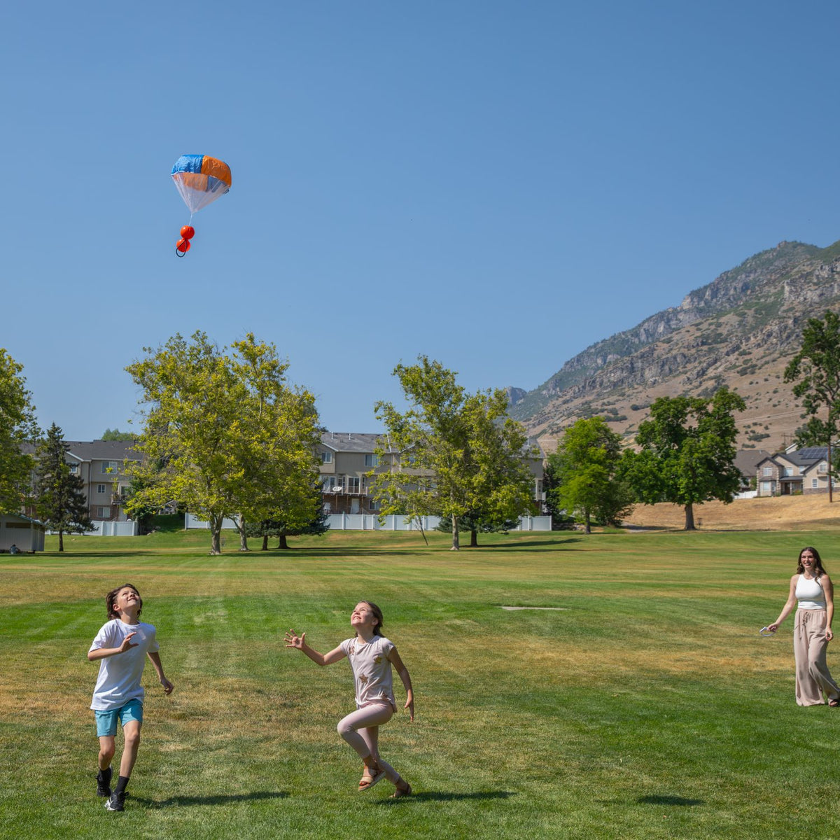 Two children chasing a ParaShoot. The parachute is drifting slowly back to earth against a blue sky, capturing the visual excitement of the slow-fall mechanism.