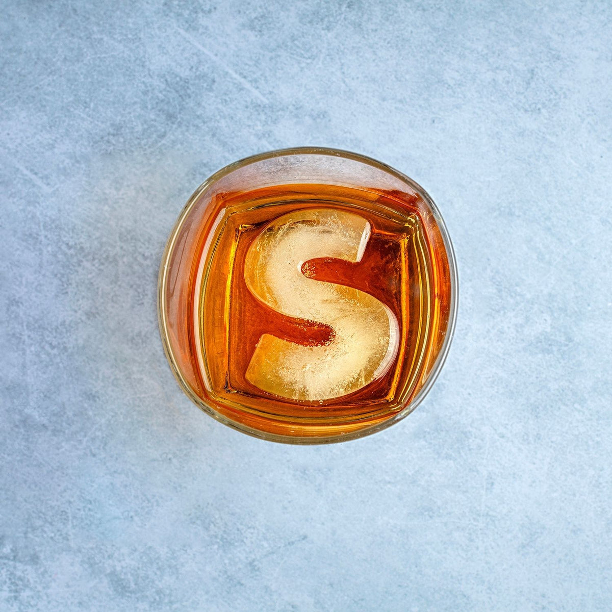 A single, large ice letter S resting in a glass of amber liquid, showcasing its slow-melting properties and unique personalised look.