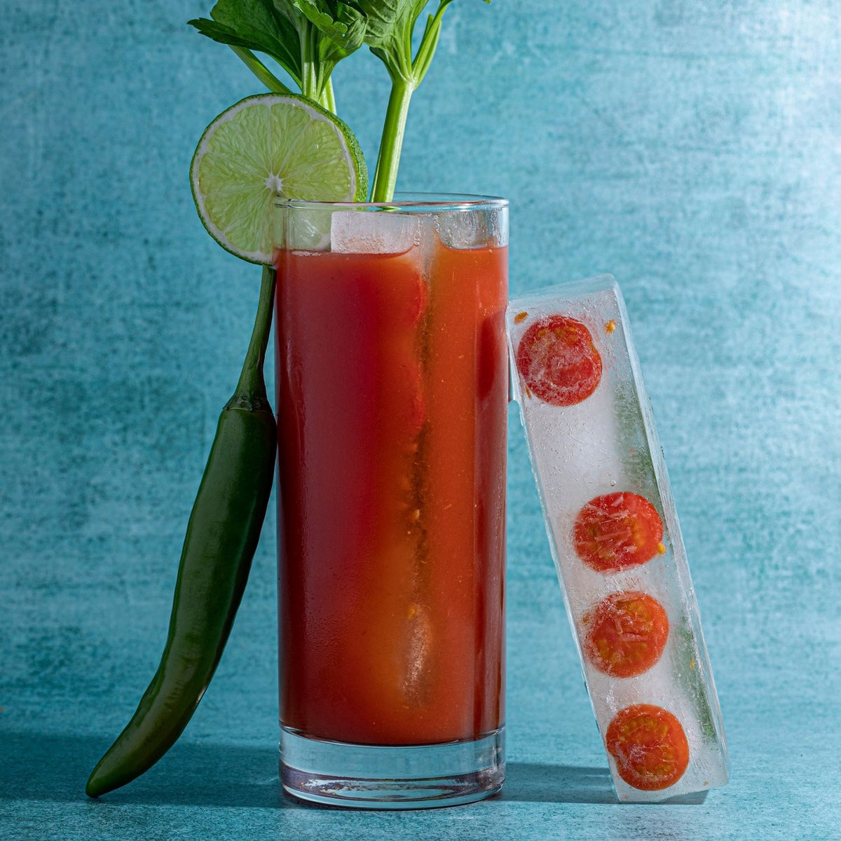 Glass of red Bloody Mary cocktail with lime, pepper, and tomato ice cubes on a blue background