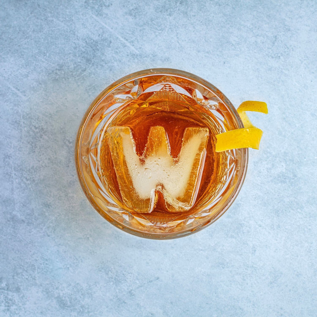A clear ice letter W being popped out of a flexible ice tray mould, showing the sharp lines and bold design of the slow-melting shape in a whisky glass.