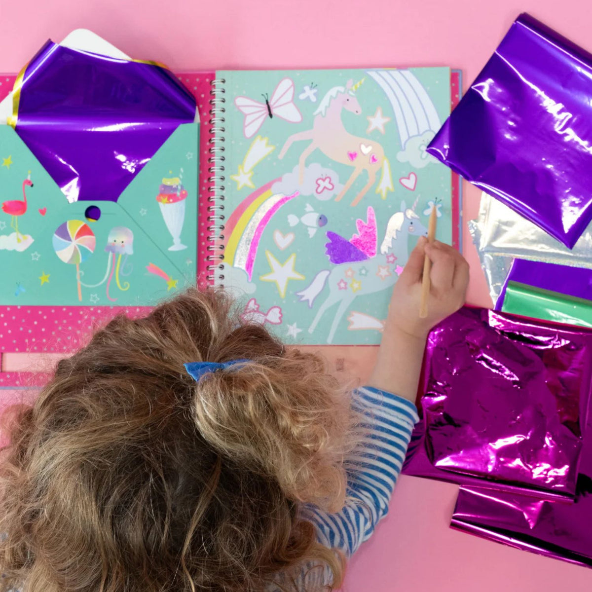 A child using the wooden stylus to rub a holographic foil sheet onto a star-shaped sticker section of a unicorn illustration.