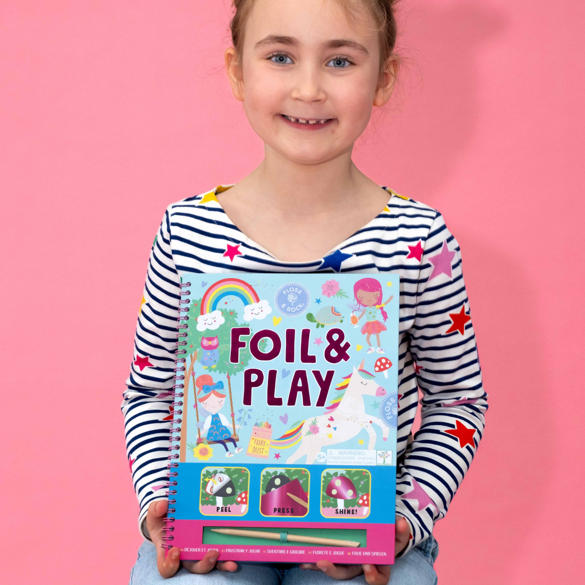 a girl holding A spiral bound book titled 'FOIL & PLAY' with illustrations of fairies and rainbow. It includes a wooden stylus and color foiling sheets.
