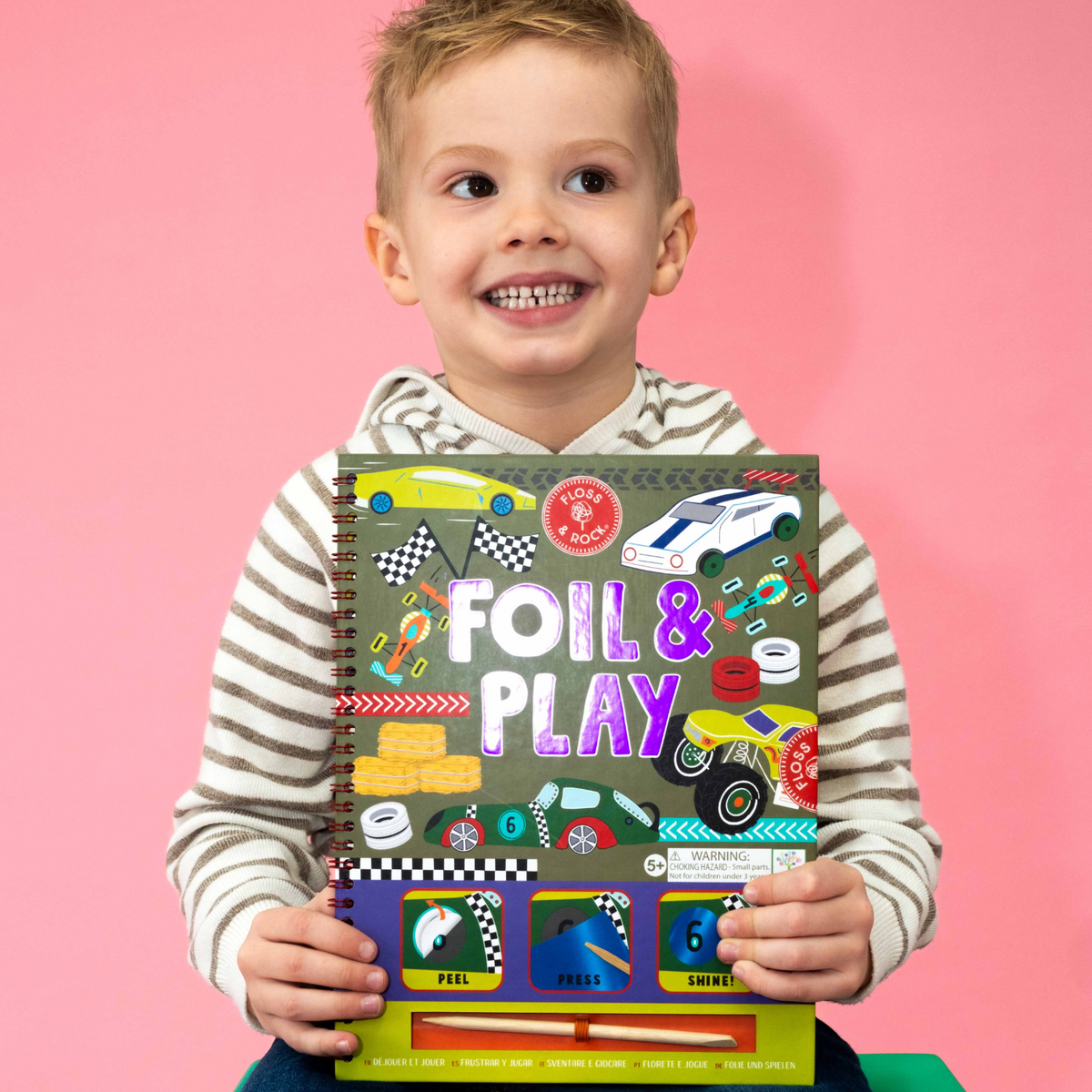 A boy holding A spiral bound book titled 'FOIL & PLAY' with colorful illustrated pages and various car designs.