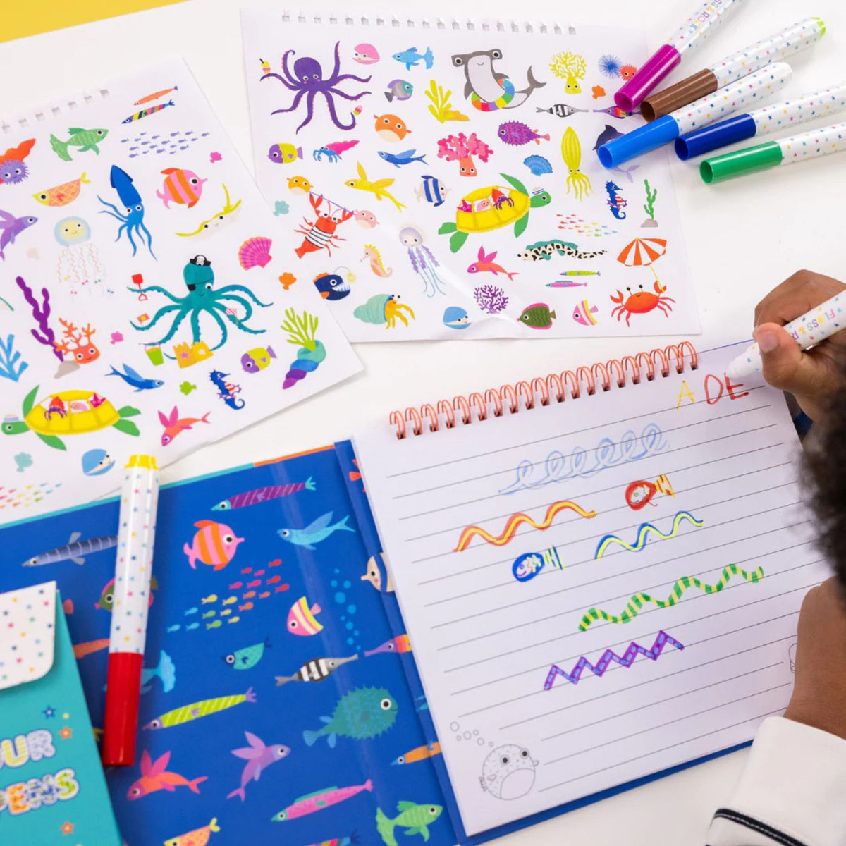 An open view of the activity pad showing a mix of a whimsical doodle page, a lined writing page, and a sheet of colourful themed stickers.