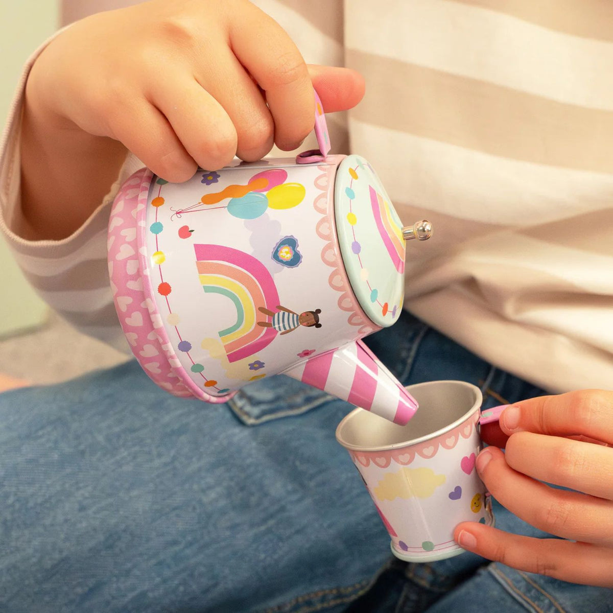A child’s hand tilting the teapot over a teacup, highlighting the wind-up key on the bottom that activates the "It's a Small World" melody.