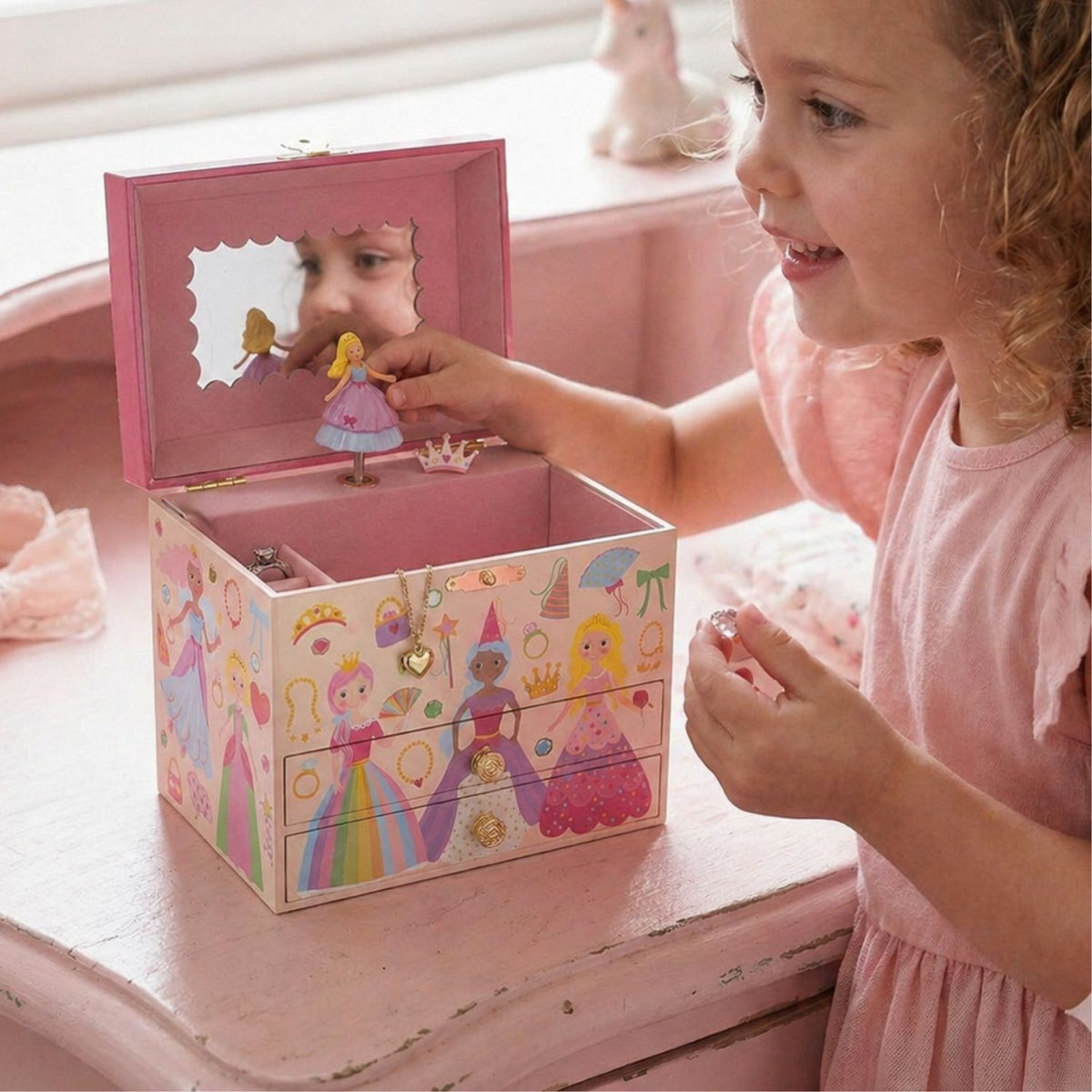 Child playing with the shimmering princess jewellery box sitting on a pink dresser, reflecting light off the metallic foil accents in a brightly decorated child's bedroom.
