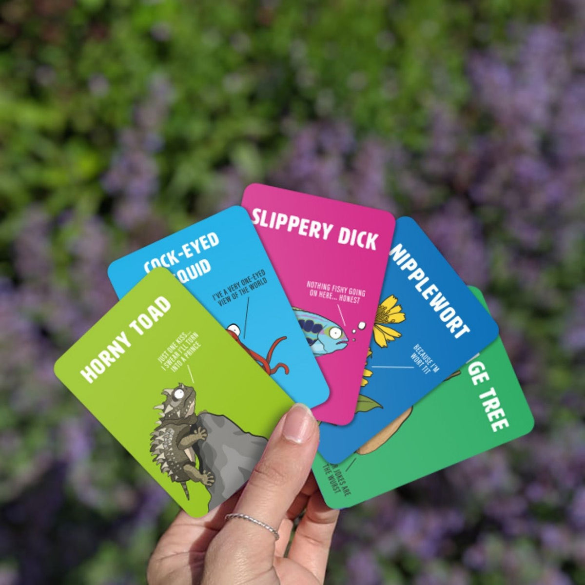 Hand holding four colorful cards with cartoon illustrations and text against a blurred natural background