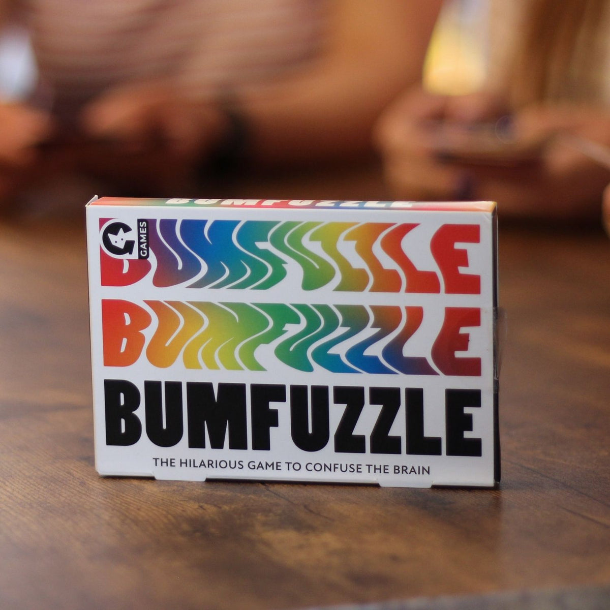 Box of BUMFUZZLE game on a wooden surface with blurred people in the background