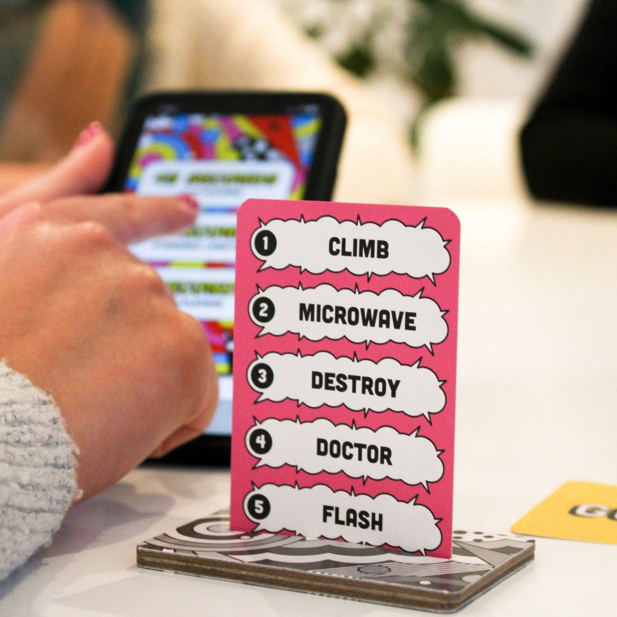 Card with numbered actions like 'Climb', 'Microwave', etc., held by a person with a phone in the background.