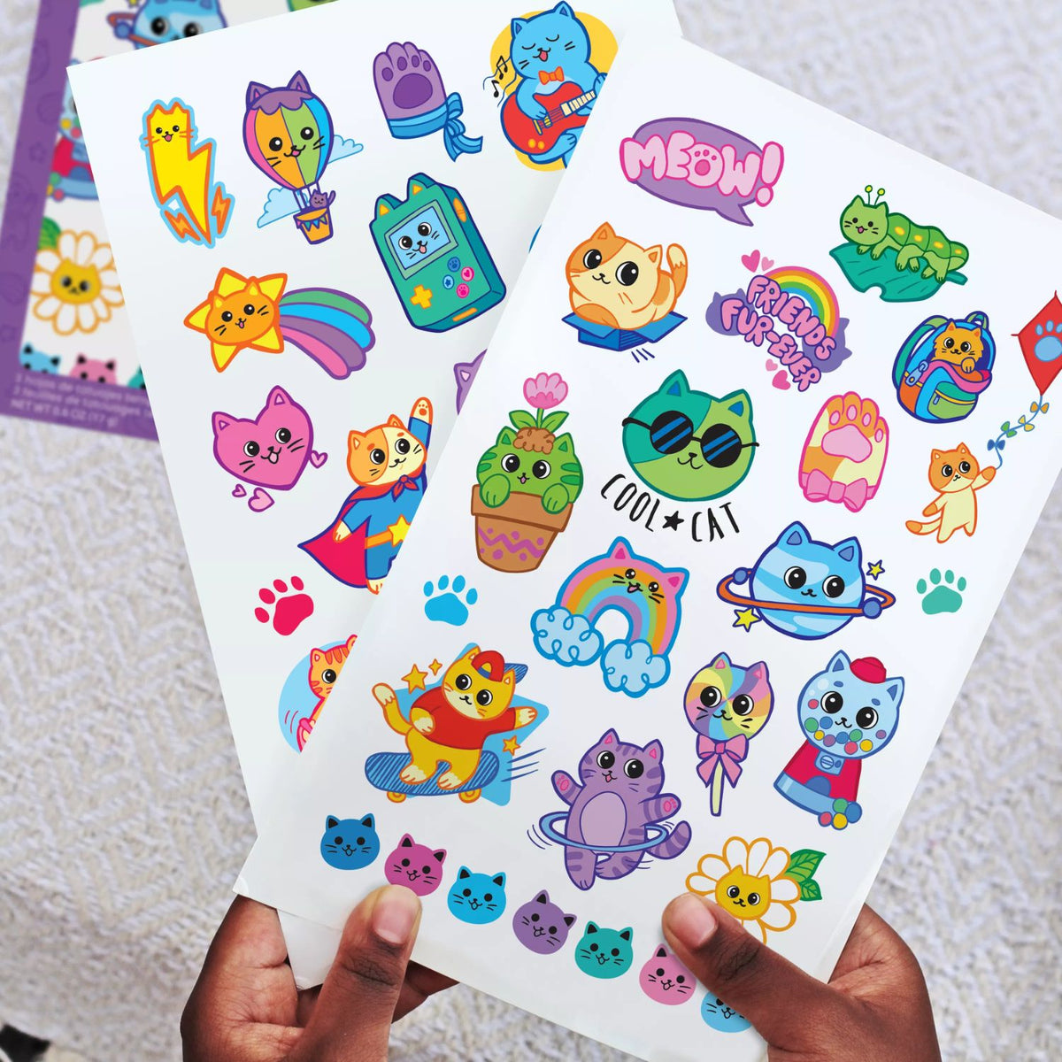 Sheet of colorful cat-themed temporary tattoos held by a hand on a textured surface.