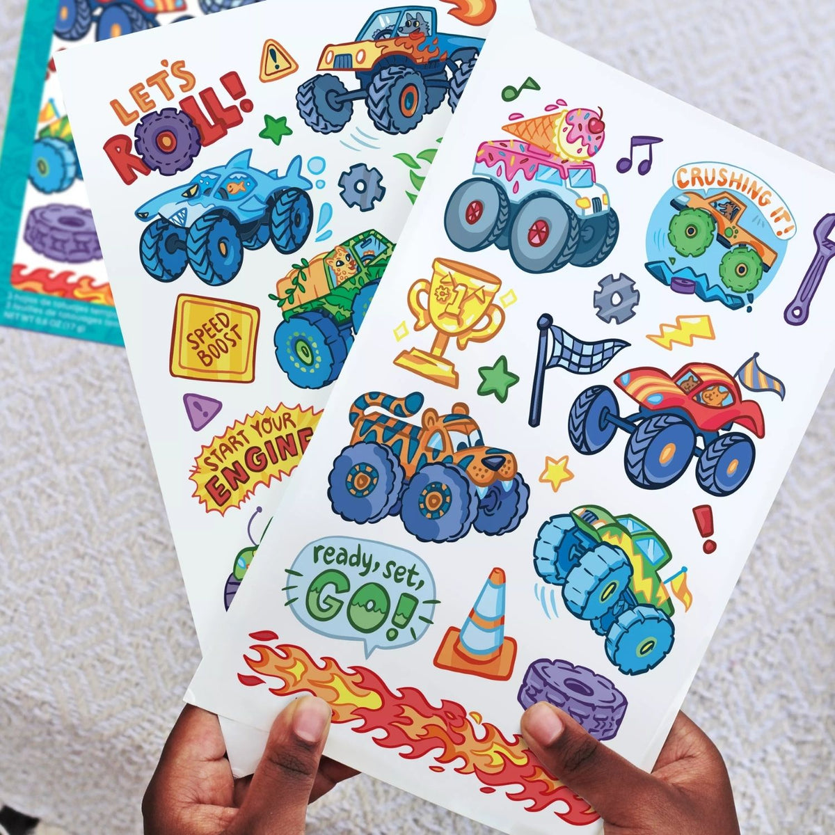 Colorful monster truck-themed stickers held by a hand on a textured surface.